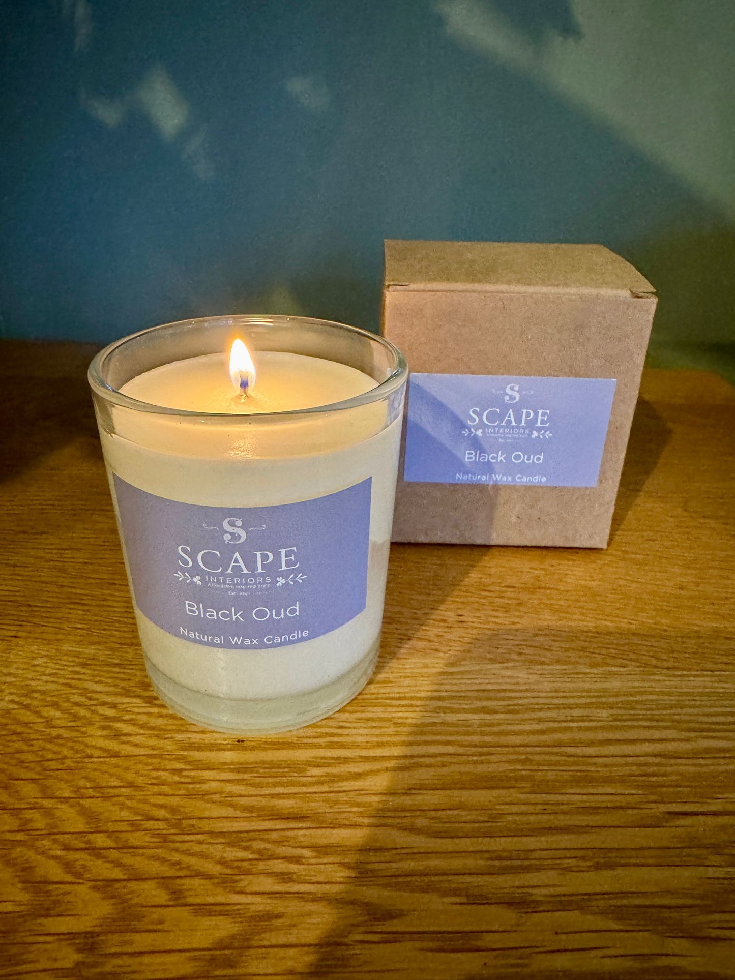 Candle labeled 'Scape' with a box in the background on a wooden surface. Back Oud Natural Wax Candle