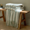 Beechwood Throw Moorland Green Folded striped blanket on a wooden bench with a candle in the background