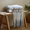 Stack of striped blankets on a wooden stool with a plant in the background Beechwood Throw Sky Blue 