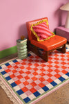 Orange chair with a pink pillow on a checkered red, blue, and white rug against a pink wall.