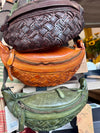 Three woven leather waist bags in brown, orange, and green on a table.